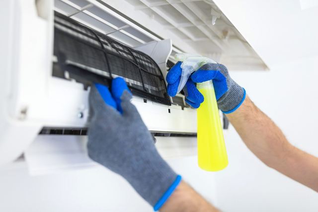Gloved hands spraying cleaner on the filter of an open wall mounted mini split