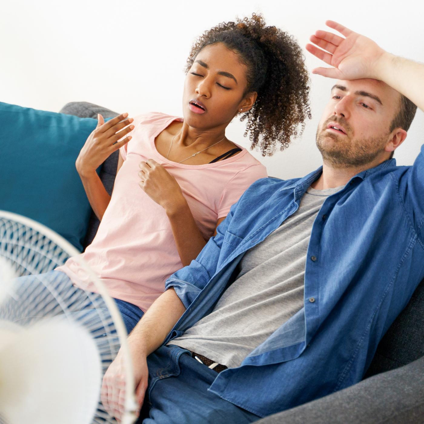 Couple sitting on a gray sofa looking overheated, woman fanning herself by hand and man wiping his forehead near an electric fan