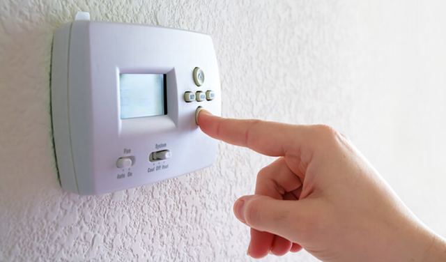 Finger pressing a button on a white digital thermostat mounted on a textured wall