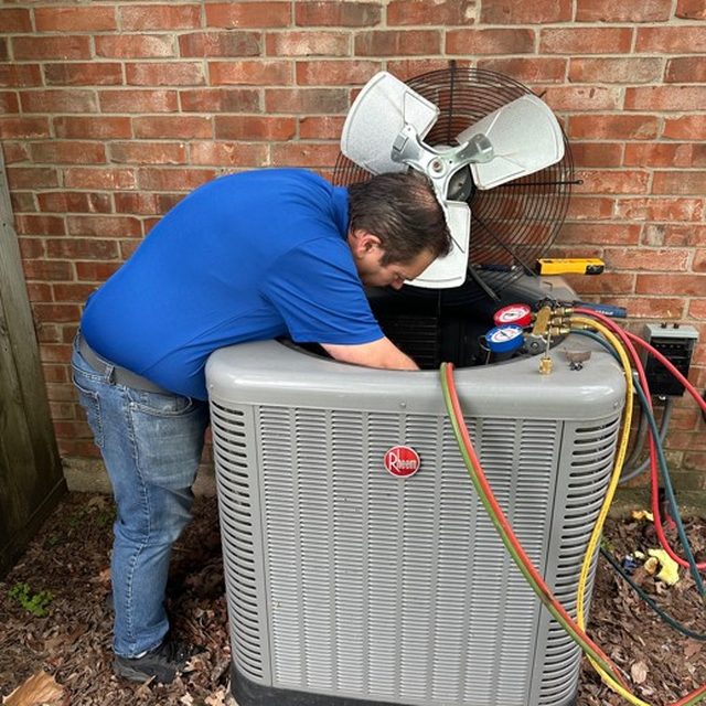 HMS owner Gary Hays leaning into an open Rheem outdoor condenser with service gauges attached, working beside a brick home