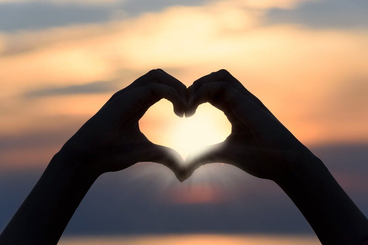 Two hands forming a heart shape, silhouetted against a sunset sky
