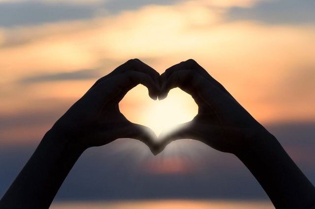 Two hands forming a heart shape, silhouetted against a sunset sky