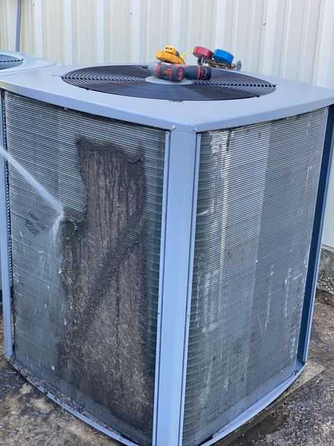 Outdoor condenser coil matted with debris on one side while an HMS technician rinses it clean