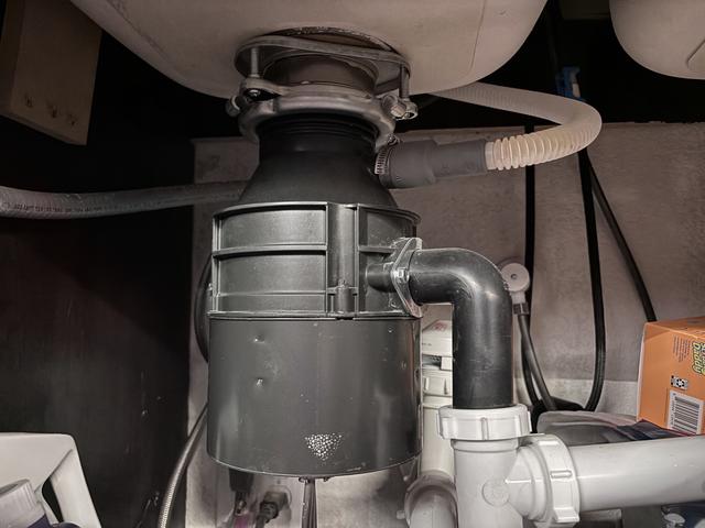 Garbage disposal mounted under a kitchen sink with its metal discharge elbow tied into a white PVC drain trap