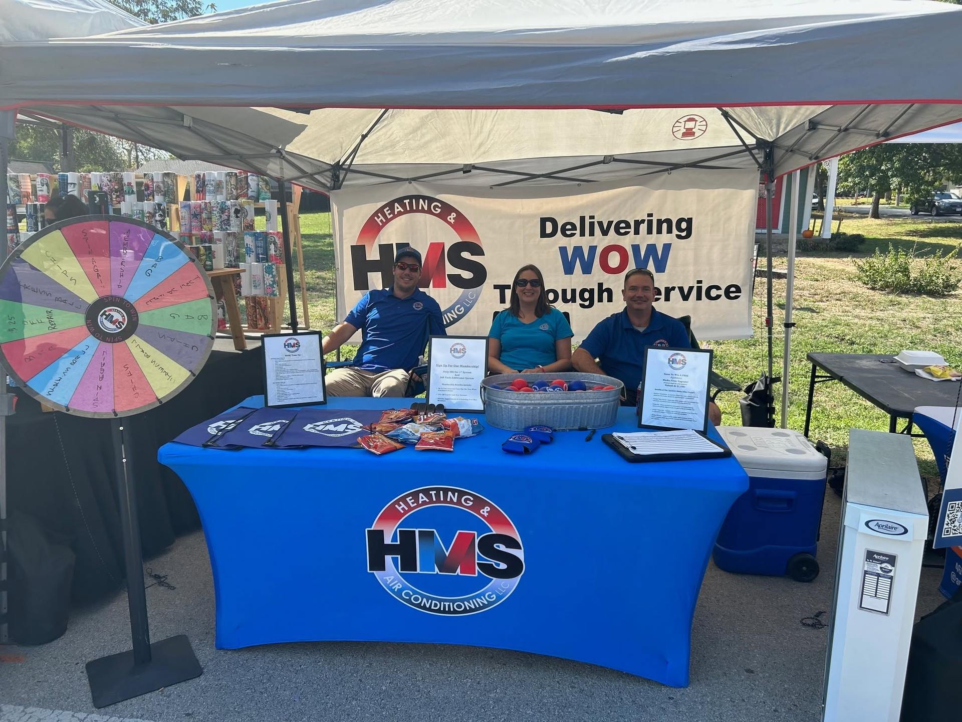 Three HMS team members at a community event booth under a Delivering WOW Through Service banner