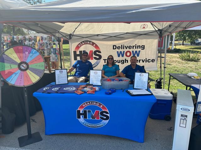 Three HMS staff at a community event booth under a canopy with a Delivering WOW Through Service banner