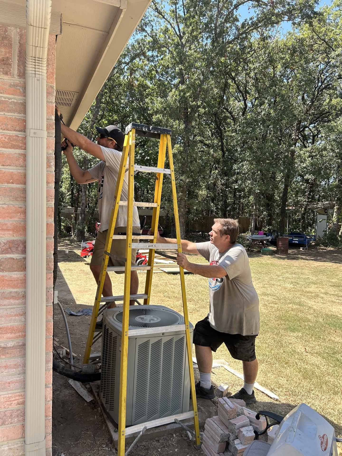 Two HMS technicians replacing an outdoor condenser beside a brick home, one working from a ladder