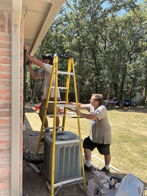 Two HMS technicians working beside a brick home, one on a stepladder at the eave above the outdoor condenser