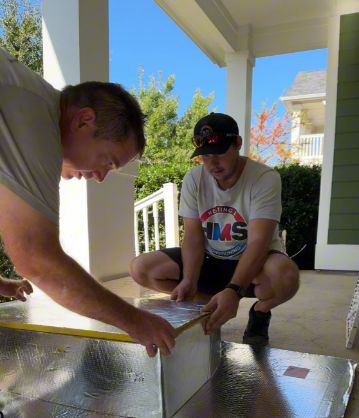 Two HMS technicians measuring and cutting duct board on a covered porch