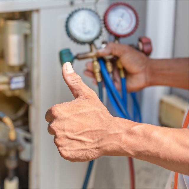 HVAC technician giving a thumbs up while holding refrigerant manifold gauges beside an outdoor unit