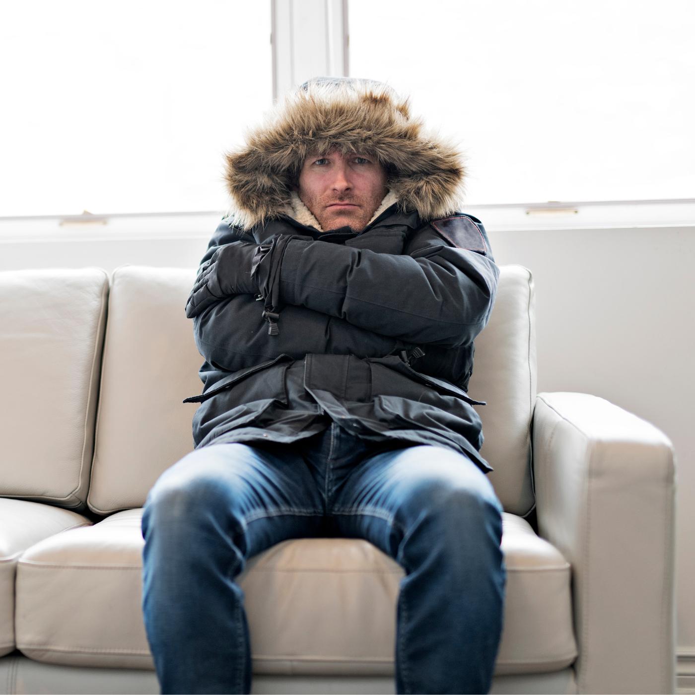 Man sitting on a cream sofa indoors wearing a hooded winter parka and gloves with arms crossed, looking cold