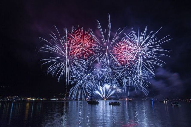 Red, white and blue fireworks bursting over a harbor at night, reflected in the water