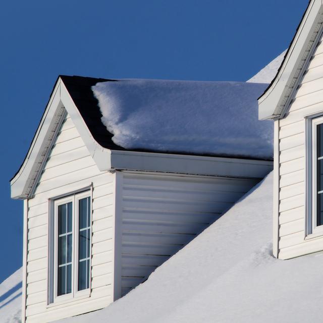 Snow-covered roof with white-sided gabled dormer windows under a clear deep blue winter sky