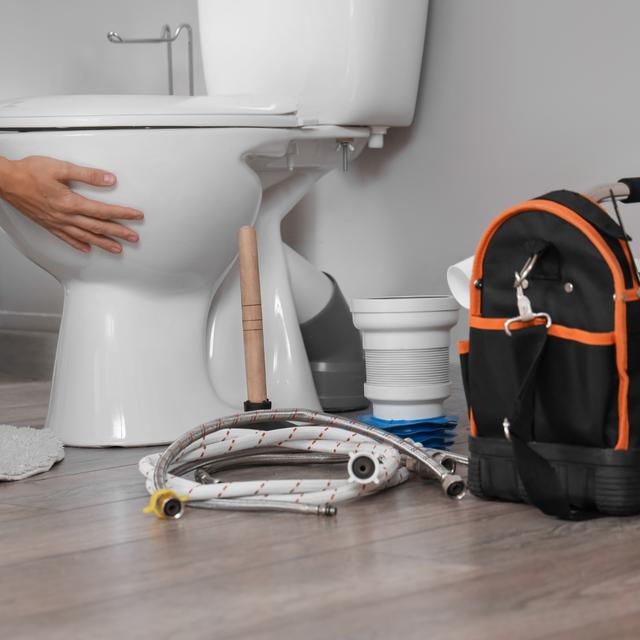 Plumber setting a new toilet in place with supply hoses, a plunger, and a tool bag on the floor
