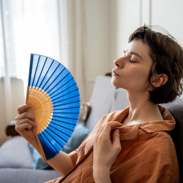 Woman with eyes closed leaning back on a gray sofa, fanning herself with a blue hand fan and pulling at her shirt collar