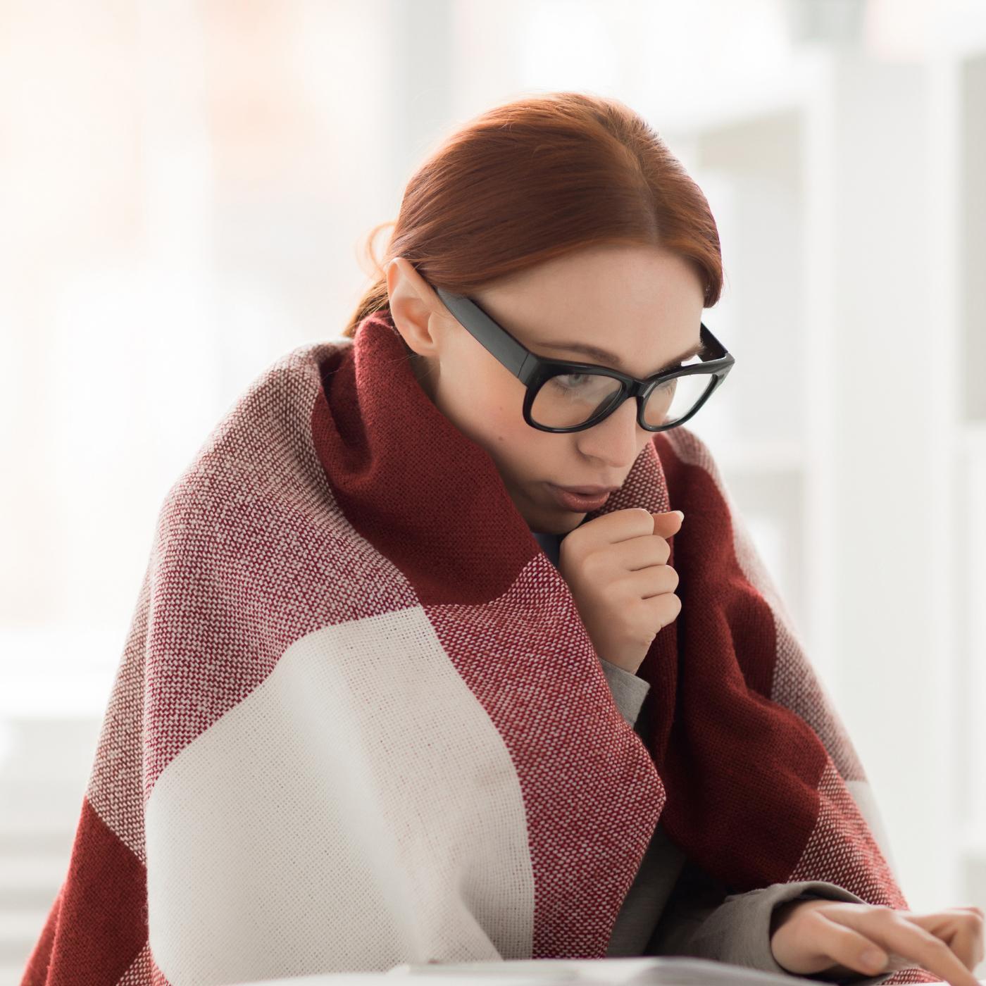 Woman in glasses wrapped in a red plaid blanket, blowing on her hand to warm it while sitting at a table