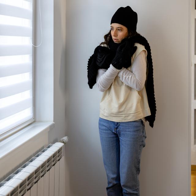 Woman in a knit hat, scarf and gloves hugging herself indoors beside a window